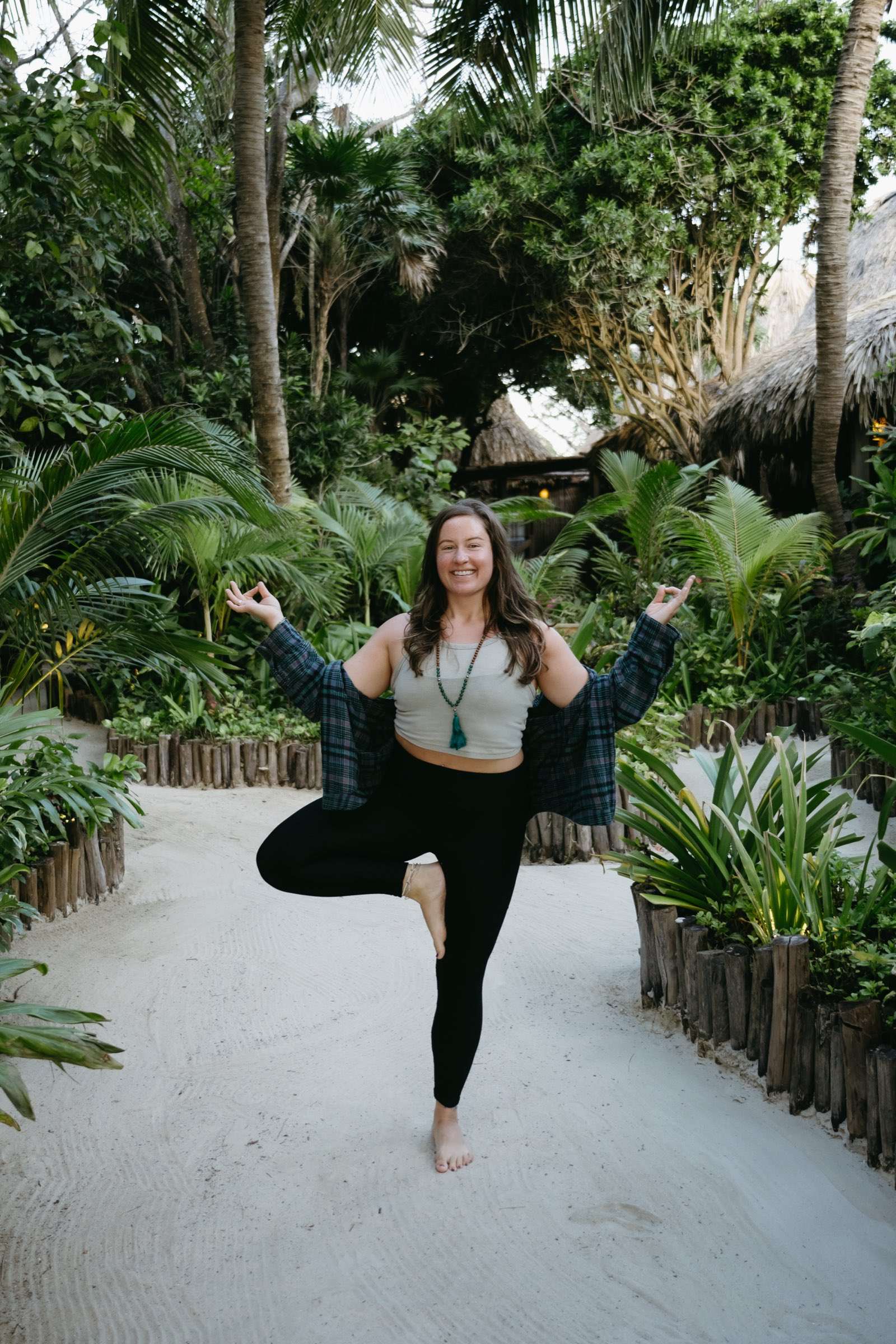 Megan smiling in tree pose on a sandy, palm-lined path